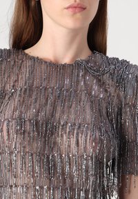 Sheer top featuring vertical rows of elongated beads in silver and transparent colors. High neckline with detailed texture and layered accents.
