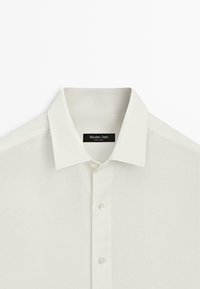 Biała koszula z kołnierzykiem w szpic i trzema widocznymi guzikami, z widoczną metką marki Massimo Dutti wewnątrz kołnierza.