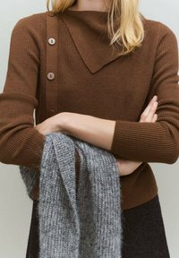Pull en tricot marron côtelé avec un col rabattu et trois boutons marron sur le côté. Écharpe en tricot grise tenue devant.