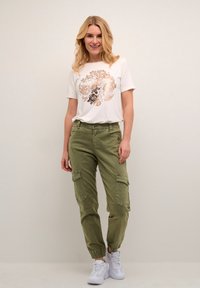 T-shirt blanc avec un imprimé floral en or rose et noir, associé à un pantalon cargo vert olive avec poches et poignets élastiques, porté avec des baskets blanches.