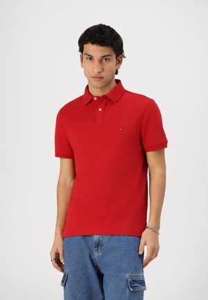 Ung mann som har på seg en rød kortermet polo og blå cargobukser, står foran en ensfarget lys bakgrunn.