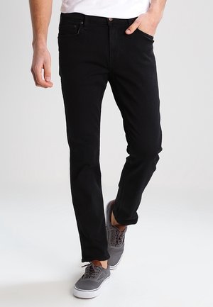 Jeans de mezclilla negra con un ajuste slim, que cuentan con cinco bolsillos, cierre de botón y detalles de costura mínimos. Llevados con zapatillas grises.
