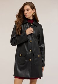 Kobieta z długimi włosami, nosząca duże złote kolczyki oraz czarny skórzany płaszcz z złotymi guzikami na bordowej sukience.