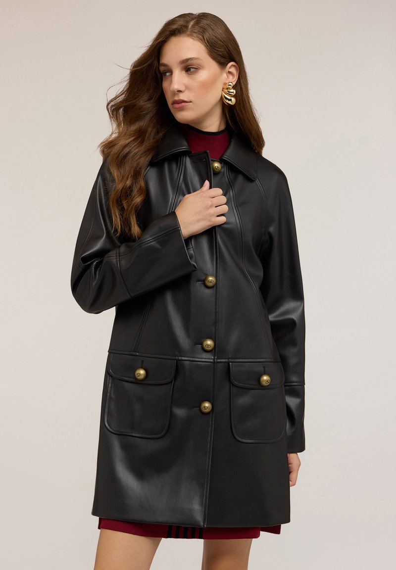 Kobieta z długimi włosami, nosząca duże złote kolczyki oraz czarny skórzany płaszcz z złotymi guzikami na bordowej sukience.