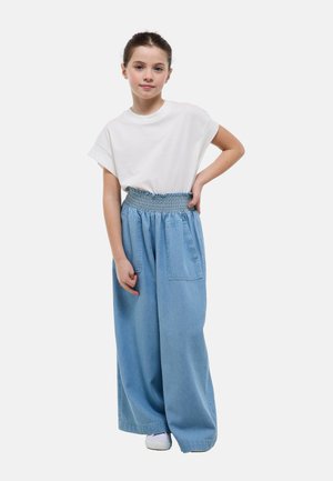 Jeune fille aux cheveux attachés portant un haut blanc à manches courtes et un pantalon bleu clair à jambes larges avec de grandes poches avant.