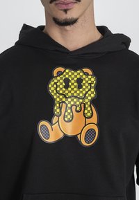 Felpa nera con cappuccio che presenta una grafica di un orso in stile cartoon, in arancione e giallo, con un design che gocciola e motivi testurizzati sui piedi.