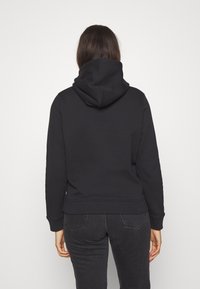 Mujer vestida con una sudadera negra y jeans oscuros, de pie de espaldas con las manos relajadas a los lados, contra un fondo liso.