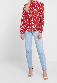 Blouse rouge à manches longues avec imprimé floral, col montant et poignets élastiques, associée à un jean en denim bleu clair déchiré et des talons blancs.