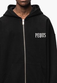 Sweat à capuche noir zippé en tissu doux, avec une poche avant et un logo texte blanc "PEQUS" sur la partie gauche de la poitrine.