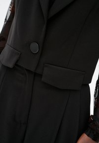 Zwarte op maat gemaakte gilet met een enkele zwarte knoop, gestructureerde revers en een riemdetail in de taille. Glad stof met strakke lijnen.