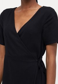 Sort slå-om-kjole med korte ærmer, dyb V-udskæring og bindedetalje ved taljen. Stoffet virker blødt og struktureret.