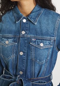 Chaqueta de mezclilla en un azul medio con cierre de botones en la parte delantera, dos bolsillos en el pecho y una cintura atada. Presenta costuras en contraste y botones plateados.