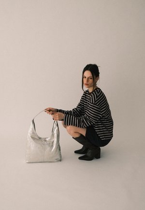 Femme accroupie portant des bottes noires et un pull rayé, tenant un grand sac à main argenté par sa poignée sur un fond uni.
