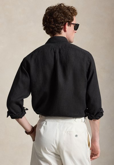 Άνδρας με σγουρά μαλλιά, που φοράει μαύρο πουκάμισο με μακριά μανίκια και διπλωμένα μανσέτα, λευκό παντελόνι, γυαλιά ηλίου και ρολόι χειρός, με την πλάτη γυρισμένη σε μπεζ φόντο.