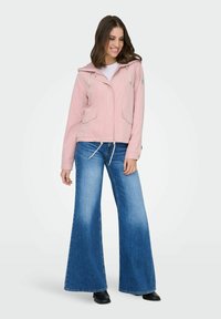 Femme souriante portant une veste à capuche rose clair, un jean bleu à jambes larges et des chaussures noires, debout devant un fond clair uni.