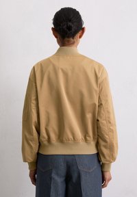 Tan bomberjack met ribgebreide kraag en manchetten, een gladde textuur, elastische tailleband en zijzakken. Achteraanzicht toont een iets oversized pasvorm.