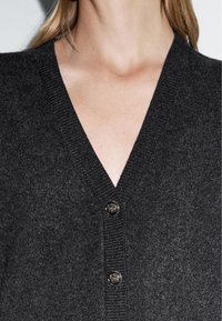 Frau trägt eine dunkelgraue, geknöpfte Strickjacke mit V-Ausschnitt auf hellem Hintergrund, die den Hals- und oberen Brustbereich zeigt.