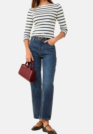Femme portant un haut à manches longues blanc et bleu marine rayé, un jean taille haute bleu, des ballerines noires, portant un petit sac à main bordeaux.