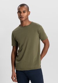 Tricou verde măsliniu cu mânecă scurtă și guler rotund, fabricat dintr-un material moale; prezintă o croială lejeră și detalii de cusătură subtile.
