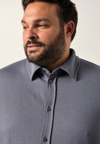 Camicia grigia con colletto a punte, texture morbida e maniche corte, con bottoni scuri e una vestibilità aderente sulle spalle.