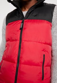 Rote Puffer-Weste mit einem schwarzen Reißverschlusskragen, ausgestattet mit einem Frontreißverschluss und kleinen seitlichen Taschen, aus glattem, glänzendem Stoff.