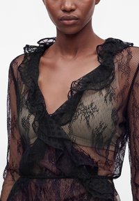Femme à la peau moyenne foncée portant un haut en dentelle noire transparente avec des motifs floraux et des bords froncés.