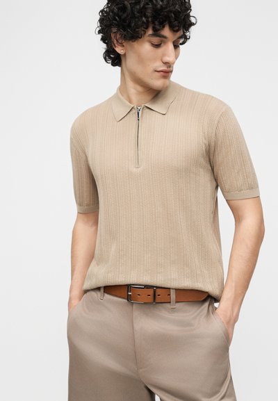 Άνδρας με σγουρά μαλλιά που φοράει μπεζ πλεκτό πόλο με κοντά μανίκια και ανοιχτό καφέ παντελόνι με αντίστοιχη ζώνη, με τα χέρια στις τσέπες.