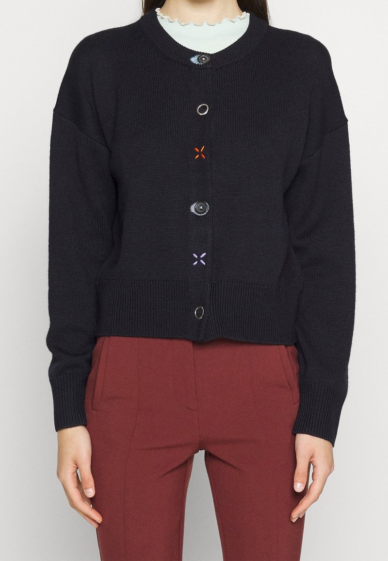 Cardigan tricoté noir avec des styles de boutons variés et des accents de fils colorés, porté par-dessus un haut bleu clair à bord festonné, associé à un pantalon taille haute rouille.