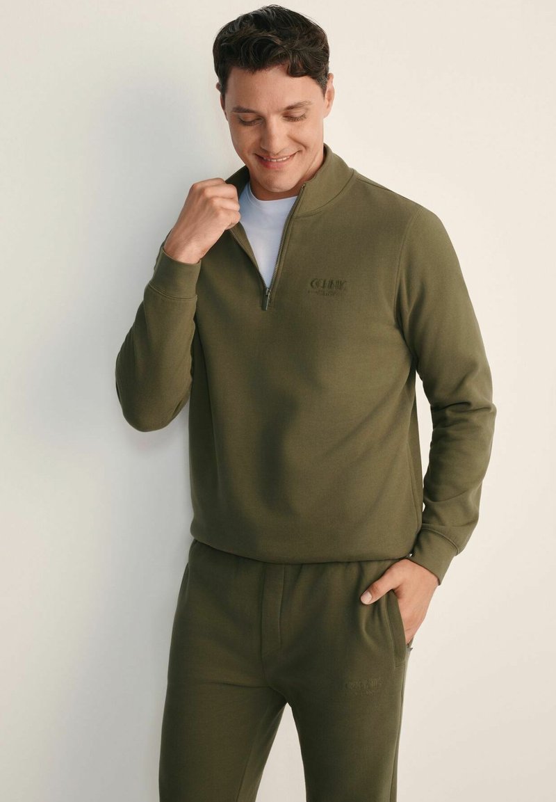 Mężczyzna poprawiający kołnierz oliwowo-zielonej bluzy z zamkiem, ubrany w pasujące spodnie, stojący na tle jednolitego jasnego tła.