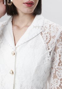 Blusa blanca de encaje con tres grandes botones de perla, intrincados patrones florales y acentos de perla en el cuello y las mangas.