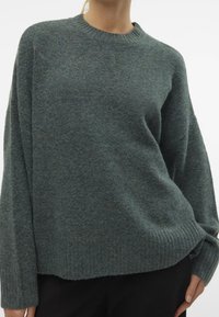 Henkilö, jolla on päällään löysä tummanharmaa villapaita ribbitettömäillä hihoilla ja helman, yhdistettynä mustiin housuihin valkoista taustaa vasten.