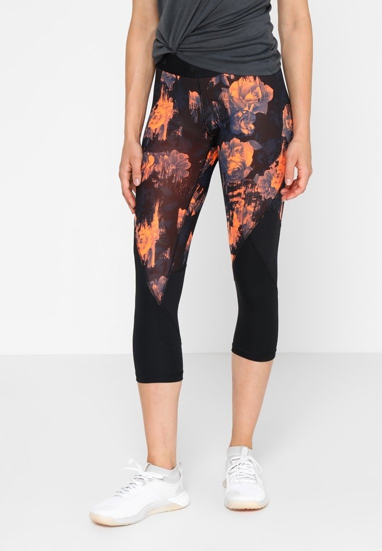 Femme portant des leggings courts à motif floral noir et orange et des chaussures de sport blanches sur un fond uni.