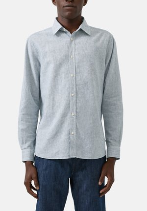 Mężczyzna w jasnoszarej koszuli z długim rękawem o delikatnej pionowej fakturze oraz ciemnoniebieskich dżinsach, stojący na jednolitym tle.