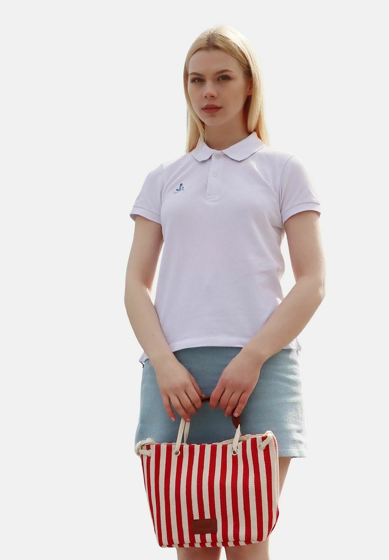 Weißes Polohemd mit kurzen Ärmeln, blauem Logo, kombiniert mit einem hellen Jeansrock. Hält eine rot-weiß gestreifte Tasche mit beigen Griffen.