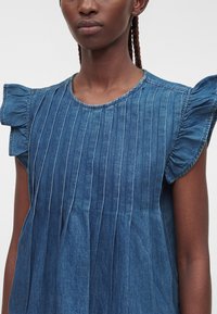 Femme portant une robe en jean bleue avec des plis verticaux et des manches courtes à volants sur un fond uni.