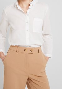 Weißes Hemd mit Knopfleiste aus leichtem Stoff, ausgestattet mit einer Brusttasche, kombiniert mit beigen, hoch taillierten Hosen, die über zwei Knöpfe verfügen.