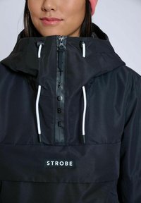 Svart huva jacka med dragkedja framtill, vita snörningar och en logotyp med namnet "STROBE." Slät textur och modern design.