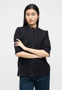 Γυναίκα με ίσια σκούρα μαλλιά που φοράει κρίκους στα αυτιά, μαύρο μπλουζάκι με δαντέλα και μπλε τζιν, με σταυρωμένα τα χέρια, μπροστά σε απλό φόντο.