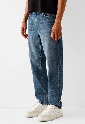 Personne portant un jean bleu ample, un haut noir et des baskets blanches, debout devant un fond clair uni.