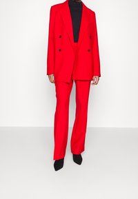 Blazer rojo de corte doble y pantalones a juego en una tela suave. Suéter negro de cuello alto debajo. Tacones negros puntiagudos completan el look.