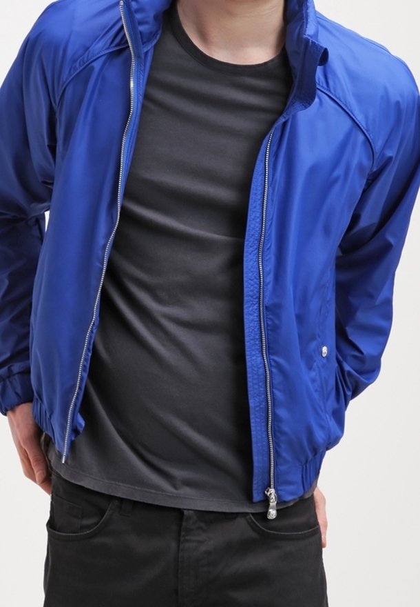 Mann trägt eine blaue Jacke mit Reißverschluss über einem schwarzen T-Shirt und schwarzen Hosen, steht mit den Händen in den Jackentaschen.