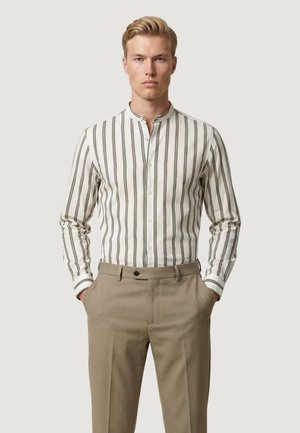 Uomo biondo con pantaloni beige e camicia bianca a righe verticali marroni, mani in tasca, sfondo neutro.