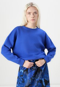 Blå hettegenser med rund hals og senkede skuldre, laget av et glatt stoff. Kombinert med et sort og blått blomstermønstrete skjørt.