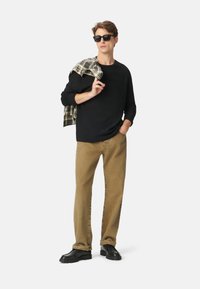 Uomo con occhiali da sole neri, maglietta a maniche lunghe nera, pantaloni beige, scarpe nere, che tiene una camicia a quadri sulle spalle, su sfondo bianco.