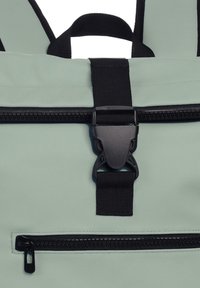 Primer plano de una mochila verde claro con un bolsillo con cremallera negra, correa negra y hebilla de plástico en la parte frontal.