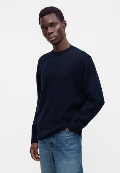 Homme portant un pull bleu marine foncé et un jean bleu, debout avec une main dans la poche devant un fond blanc uni.