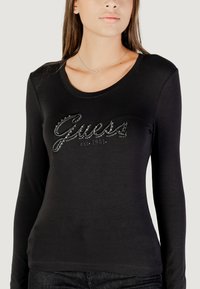 Maglietta nera a maniche lunghe con scollatura tonda e un prominente logo "Guess" in rilievo decorato con strass sul petto.