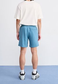 Ljus beige kortärmad skjorta, blå bomullsshorts, elastisk midja, liten bakficka, matchat med svart och vita sneakers på en blå matta.