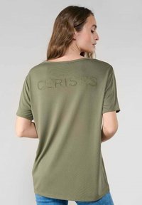 Camiseta verde oliva de tela suave, con mangas cortas y la palabra "CERISES" en letras perforadas en la parte de atrás.