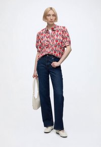 Femme aux cheveux blonds courts portant un chemisier rose à motifs à manches courtes, un jean bleu foncé, des chaussures blanches et tenant un sac à main blanc.